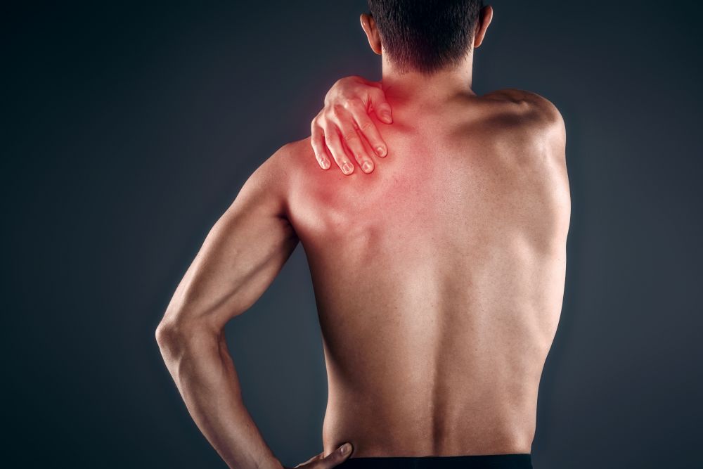 A man seen from behind clutching his upper back and shoulder, with a red-highlighted area indicating muscle pain or inflammation.