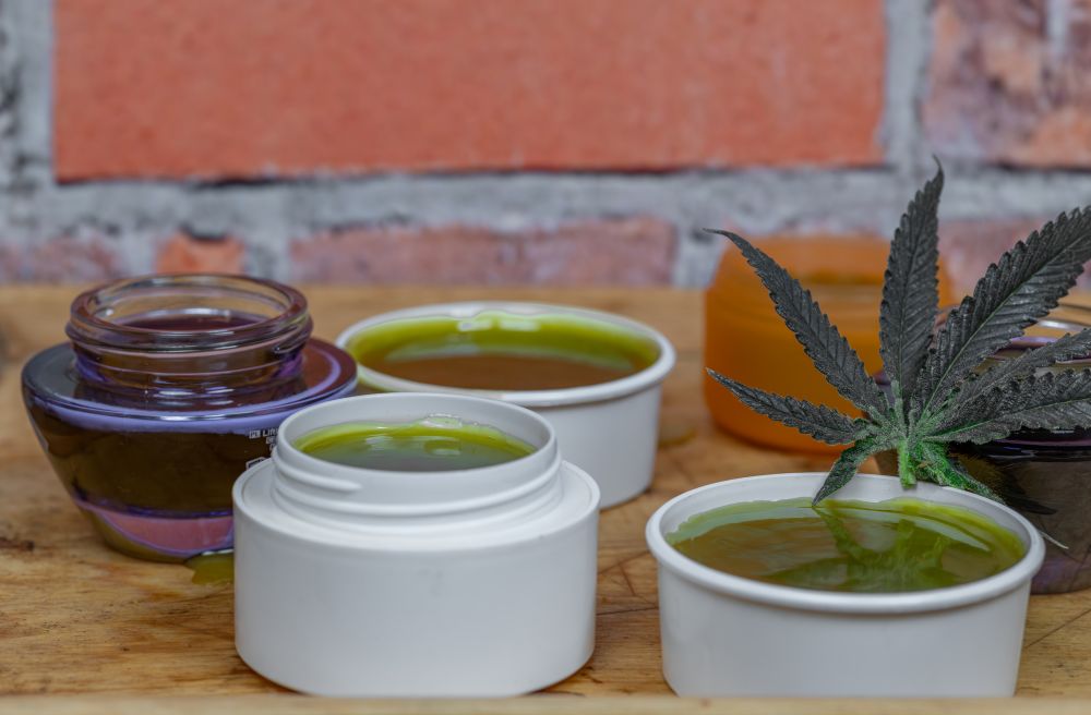 Various jars of green cannabis topicals and salves arranged on a wooden surface, with a cannabis leaf displayed beside them against a brick wall background.