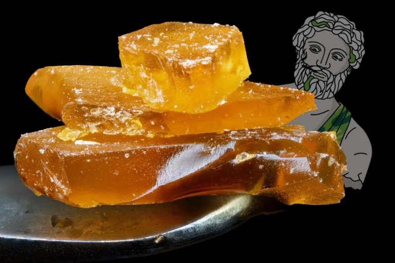 Macro shot of amber-colored cannabis shatter concentrate stacked on a dab tool, with Theo dispensary’s illustrated mascot in the background