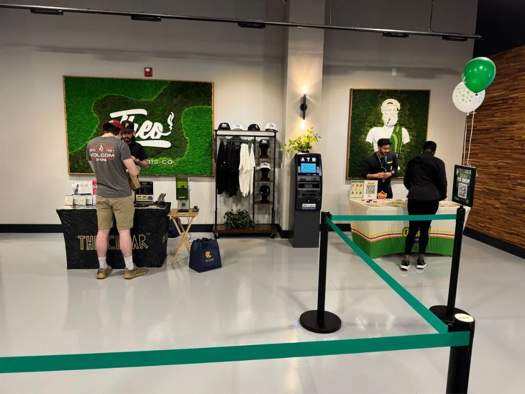 Interior of Theo Dispensary in New Jersey showing vendor booths, an ATM, branded merchandise, and staff assisting customers under a green moss wall with the Theo logo. Balloons and a relaxed atmosphere highlight the welcoming retail space.