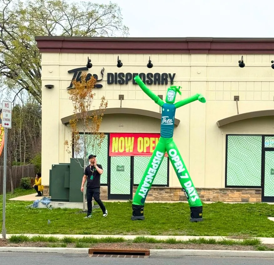 Front view of Theo Dispensary in New Jersey with a green inflatable tube man reading “OPEN 7 DAYS” and a large “NOW OPEN” sign in the window, capturing a casual street scene with a person walking by.