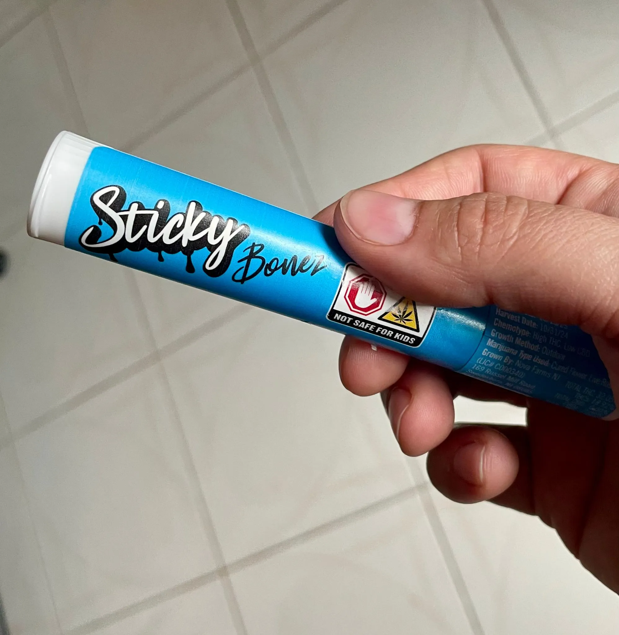 Close-up of a hand holding a blue tube labeled “Sticky Bones” — a 1g rosin infused cannabis pre-roll. The packaging features safety icons and product details.