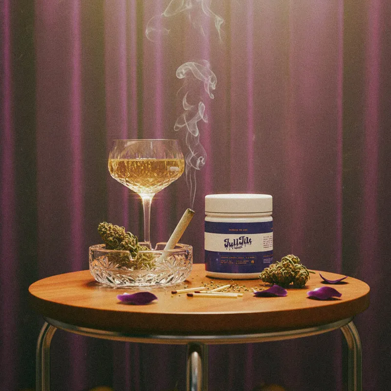 Full Tilt cannabis flower displayed with a lit joint, glass of wine, and jar on a small table under moody lighting