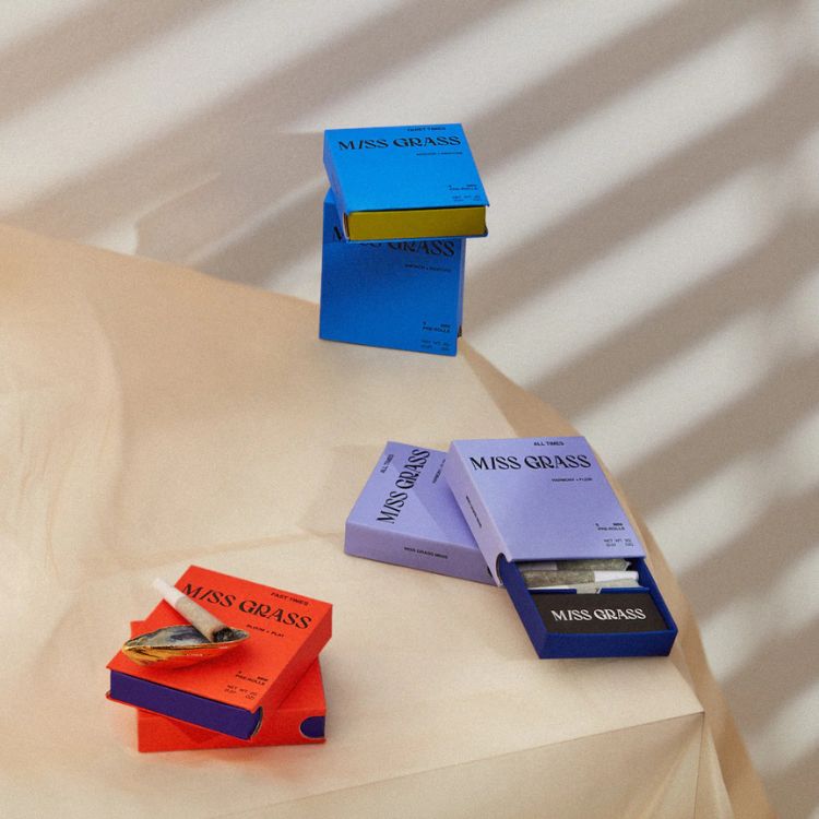 Assorted colorful Miss Grass pre-roll packs in blue, lavender, and coral placed on a beige fabric surface with diagonal shadows, showcasing a modern and lifestyle-oriented cannabis product lineup.