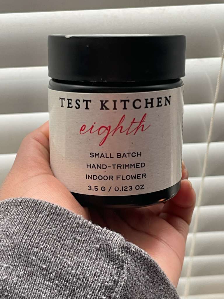 A hand holding a jar of Test Kitchen cannabis labeled “eighth – small batch, hand-trimmed indoor flower, 3.5g / 0.123 oz,” with a simple cream label and black lid, photographed against window blinds in natural light.
