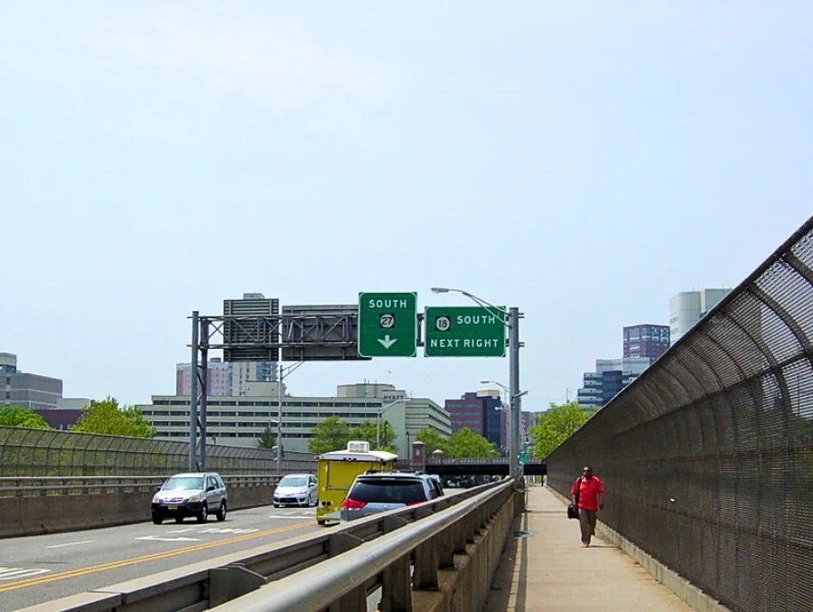 Highway view showing Route 27 South road signs in New Jersey, with cars driving toward the city skyline and a pedestrian walking along the fenced sidewalk.