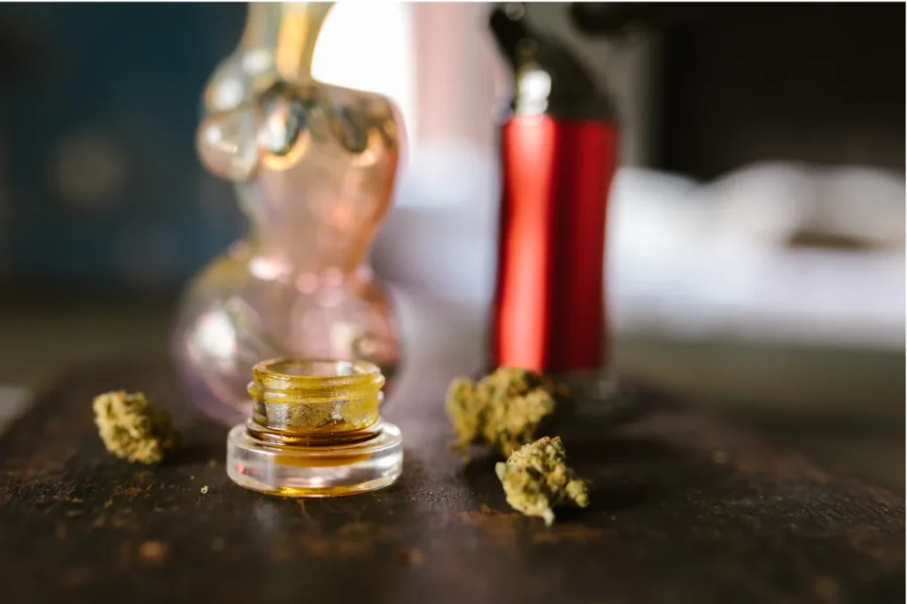 Amber cannabis concentrate in a glass jar on a dark wooden surface, with a pink glass dab rig, red butane torch, and scattered cannabis flower buds