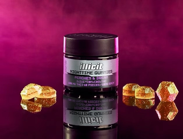Illicit Nighttime Gummies jar labeled Peaches & Dream, black cylindrical container with holographic purple label on a dark magenta background, surrounded by amber and pink faceted gummy pieces, 2:2:1 CBN:CBD:THC ratio, available at Theo dispensary in New Jersey
